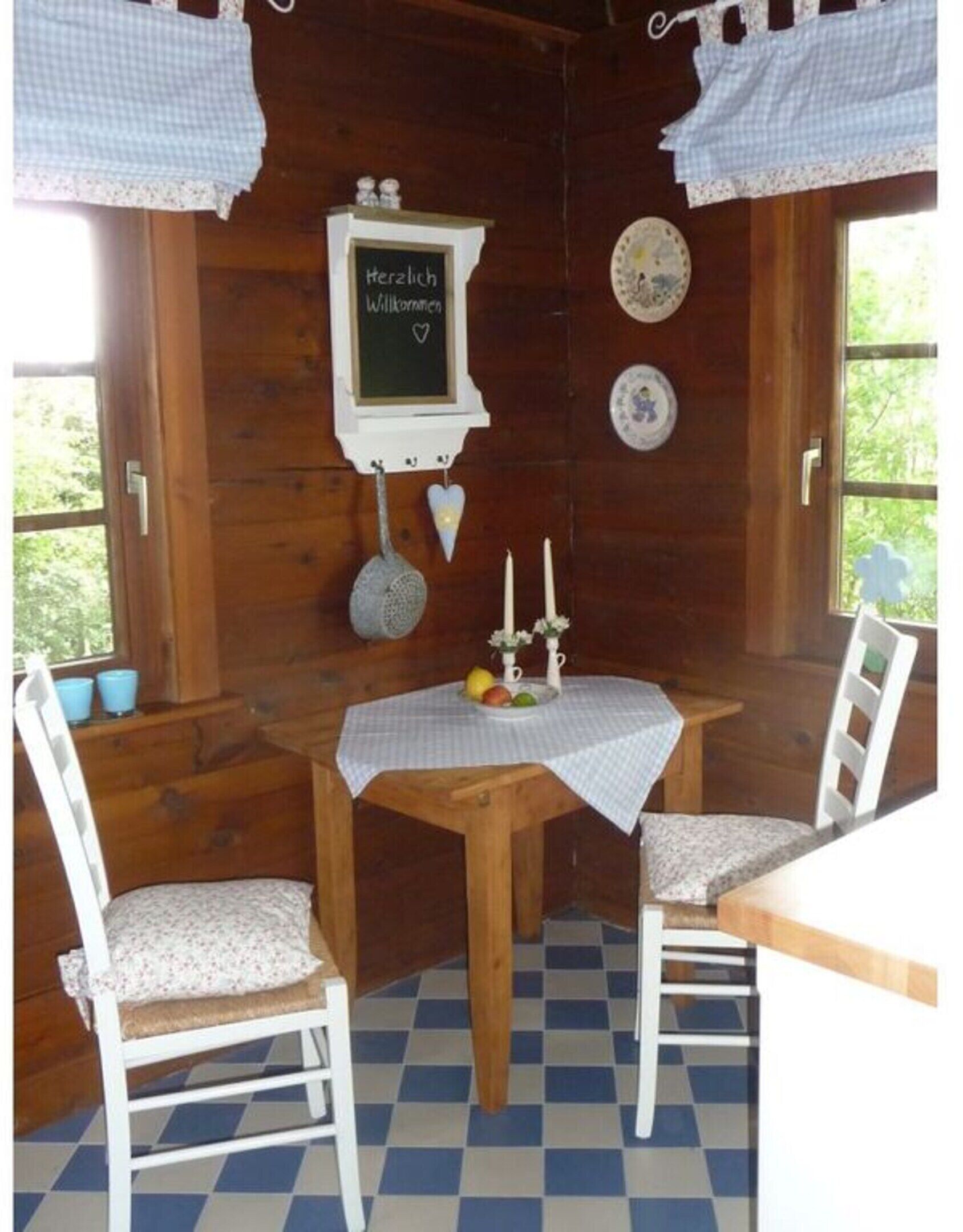Ein gemütliches Holzblockhaus-Interieur mit einem Schachbrettfußboden, einem Tisch mit Tischdecke, zwei Stühlen, einer Tafel mit einer Begrüßung und dekorativen Tellern an der Wand.