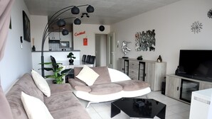 Un salon moderne avec un canapé d’angle, une table basse et un téléviseur à écran plat. On y trouve également un coin repas avec une table et des chaises, et une cuisine visible au fond.