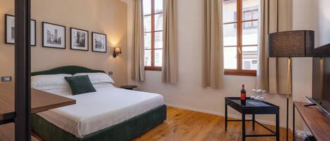 A hotel room with a bed, a small table with a bottle and glasses, a lamp, and framed pictures on the wall.