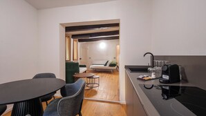 Una cocina moderna con encimera negra, estufa integrada y cafetera. Hay una zona de comedor con una mesa redonda y dos sillas. También se ve una pequeña mesa redonda con una silla al fondo.