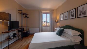 Habitación de hotel con cama, televisión, estante con vasos y cafetera, ventana con vista a edificios y cuadros en la pared.