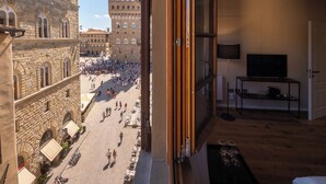 Una habitación con un amplio ventanal que da a una plaza concurrida y edificios históricos.