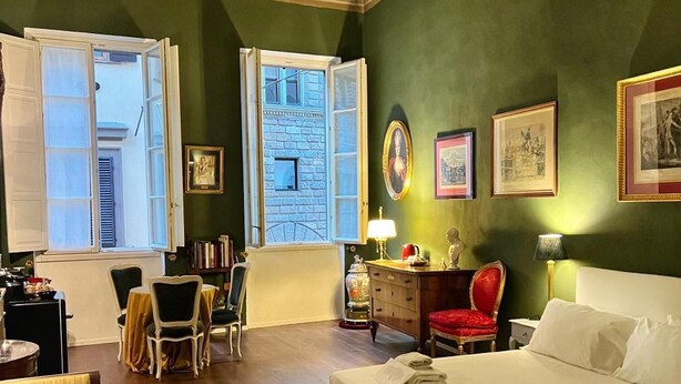 Habitación de hotel con una cama grande, un escritorio con silla, un área de comedor con sillas y vista a un edificio a través de ventanas abiertas.