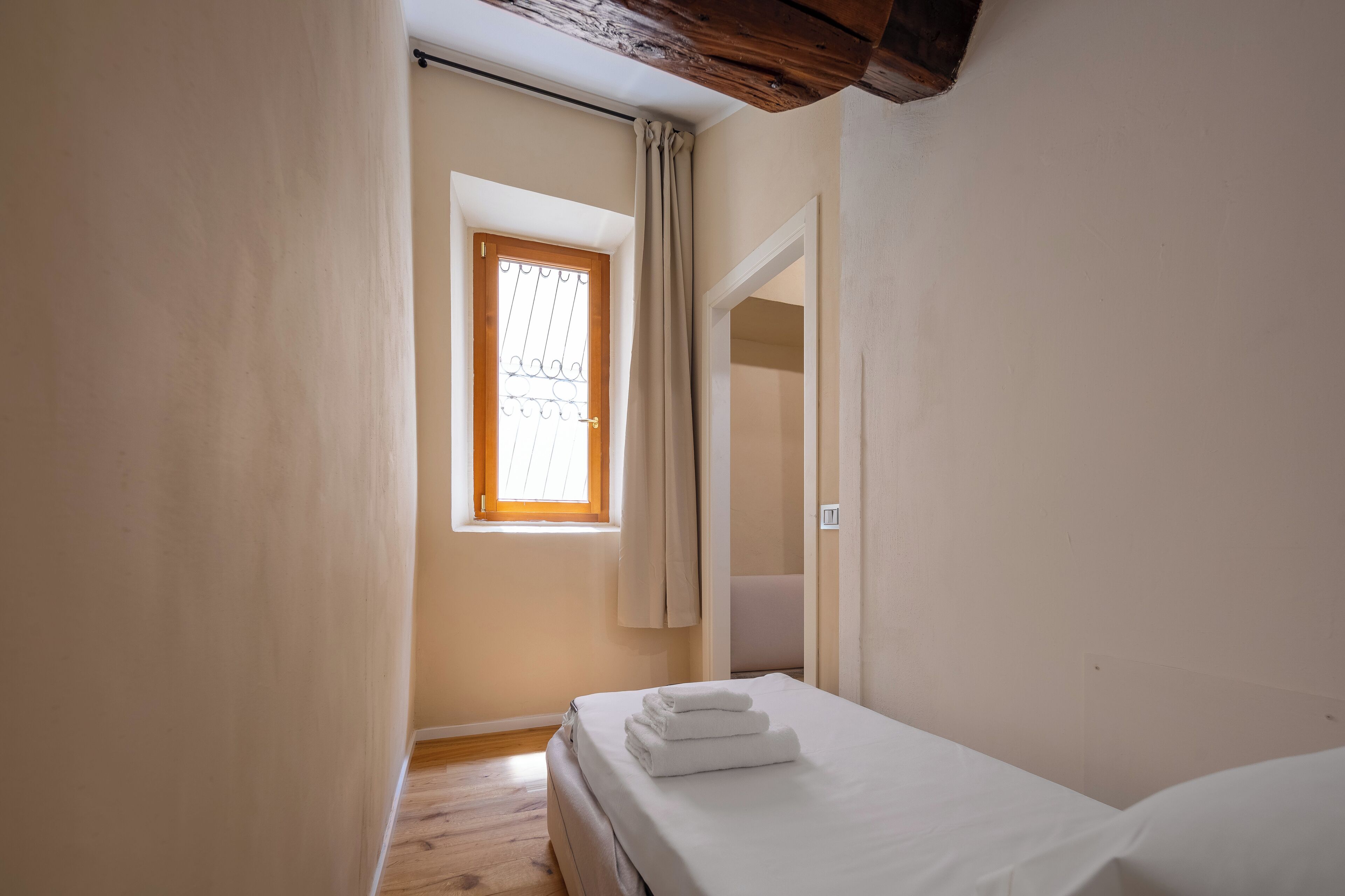 Une petite chambre bien éclairée, avec un plancher en bois, un lit simple recouvert de draps blancs, une fenêtre avec un rideau et une porte donnant sur une autre pièce.