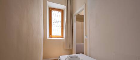 A small, well-lit bedroom with a wooden floor, a single bed with white linens, a window with a curtain, and a door leading to another room.