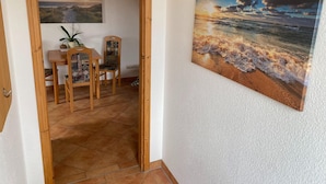 A room with a wooden door leading to a dining area with a table and chairs, a painting of a beach scene on the wall, and a staircase in the background. - Hideaway, Pruchten- Baltic Coast (Pruchten)