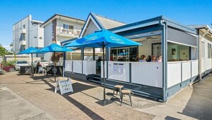 A seaside restaurant with outdoor seating, covered by blue umbrellas, and a view of the ocean. - Ocean Spray 8 - 2BR Perfect Coastal Retreat in Vibrant Coffs Harbour (Coffs Harbour)