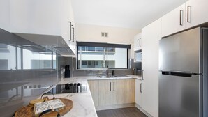 A modern kitchen with a stainless steel refrigerator, wooden cabinets, a marble countertop, and a large window providing natural light. - Ocean Spray 8 - 2BR Perfect Coastal Retreat in Vibrant Coffs Harbour (Coffs Harbour)