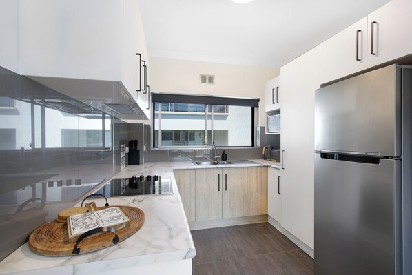 A modern kitchen with a stainless steel refrigerator, wooden cabinets, a marble countertop, and a large window providing natural light.