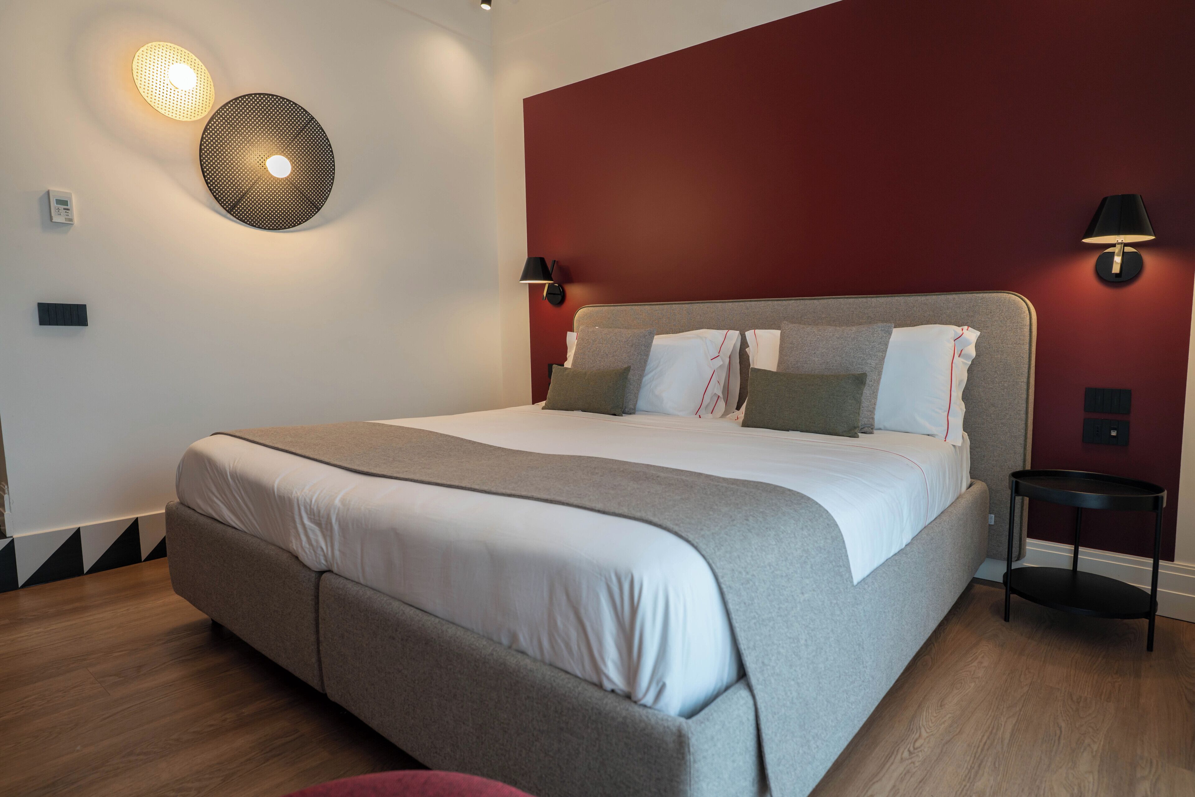 A modern hotel room with a large bed, a round wall light, a small table, and a red accent wall.