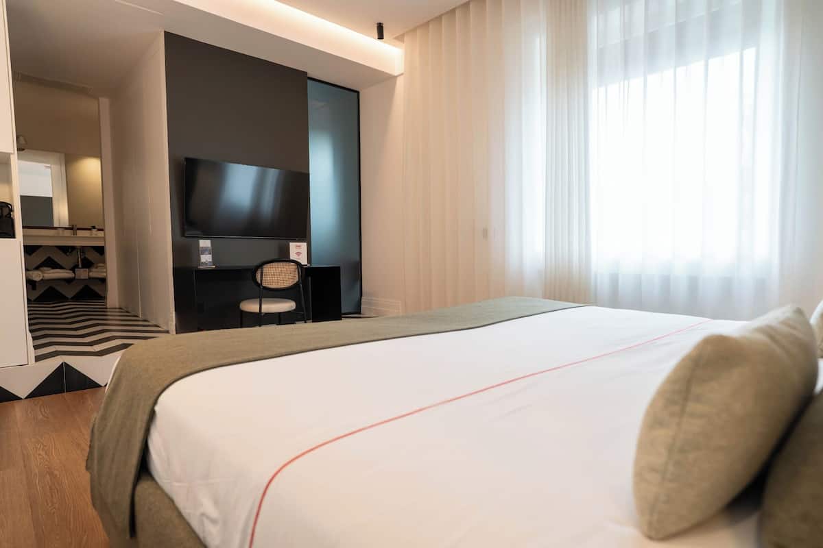 A modern hotel room with a large bed, a flat-screen TV, a desk with a chair, and a view of the outside through sheer curtains.