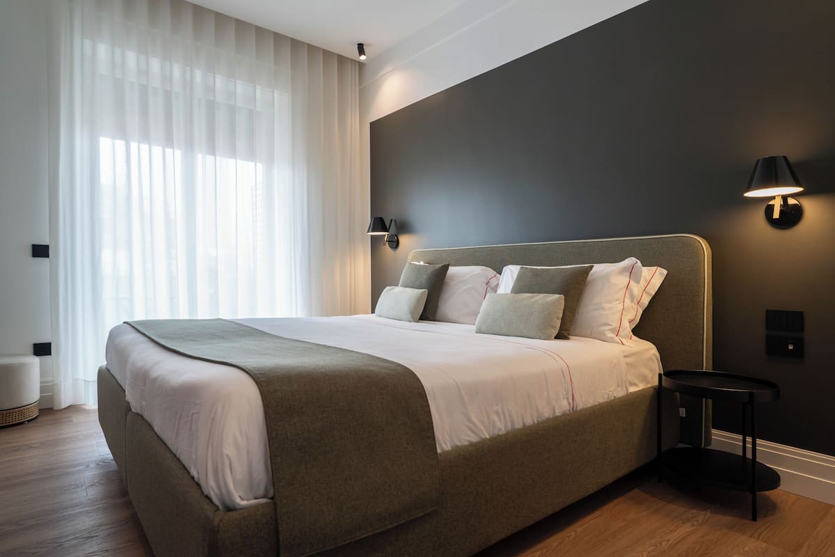 A modern bedroom with a large bed, a bedside table, a wall-mounted lamp, and a window with sheer curtains.