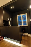 Un espace de bureau moderne avec un comptoir d’accueil de couleur sombre, un vase en verre contenant des tiges de roseau, un fauteuil de bureau noir et une fenêtre donnant sur un immeuble.