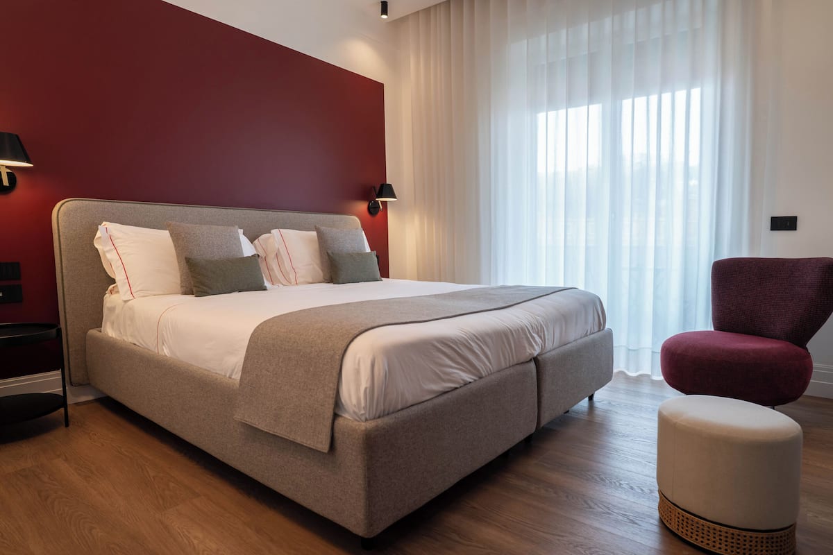 A modern bedroom with a large bed, a red accent wall, and a window with sheer curtains.