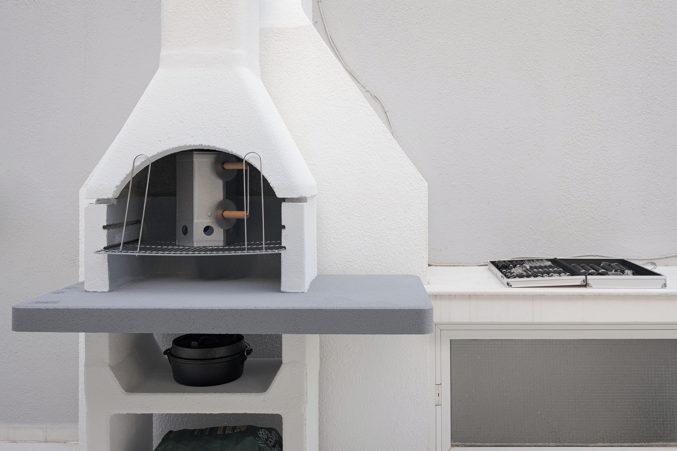 A white outdoor kitchen with a built-in oven, a shelf below holding a pot, and a grill on the right.