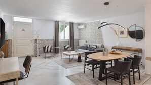 A modern living room with a sofa, coffee table, and dining area. There is a door with a sign, a window with curtains, and an air conditioning unit on the wall. - Athenian Secret Glyfada (Glyfada)