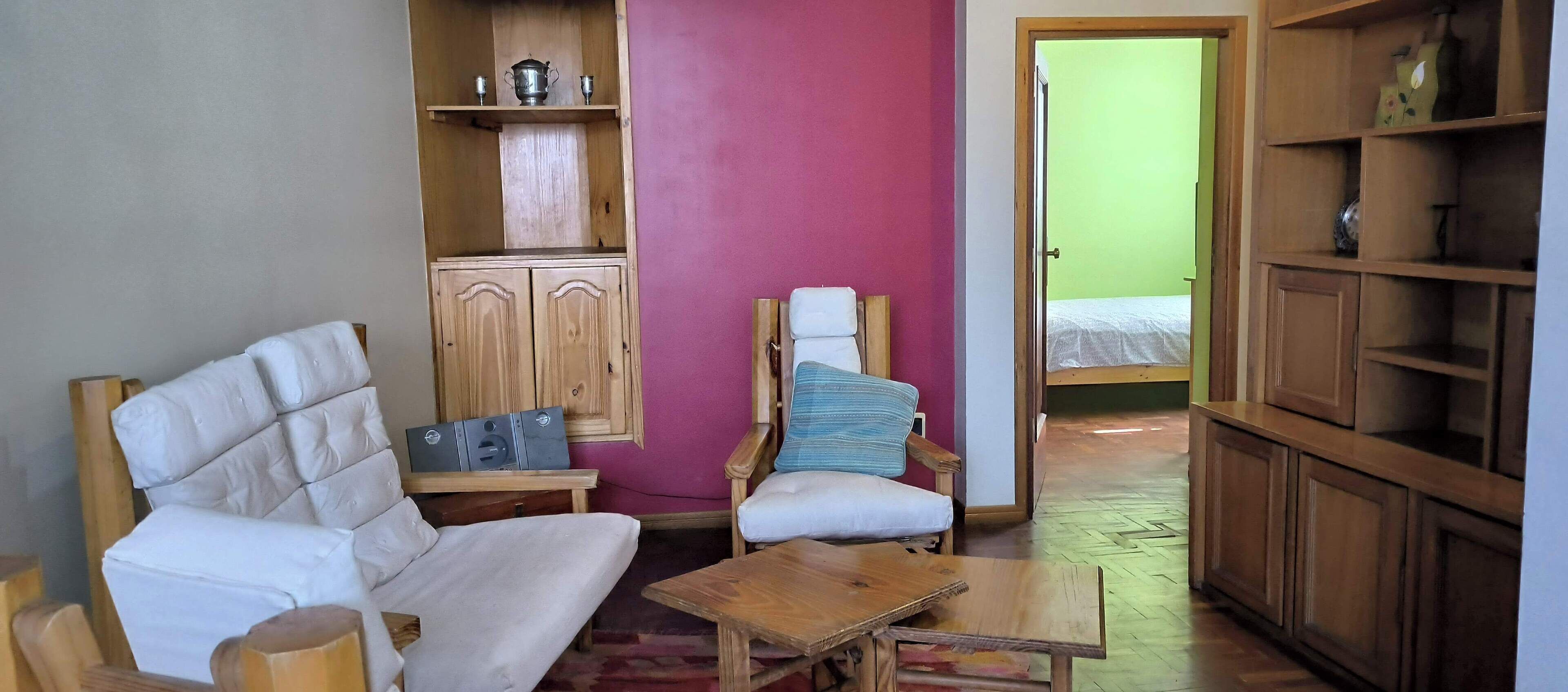 A compact living space with a white sofa, a wooden chair with a cushion, a small wooden table, and a cabinet with shelves.