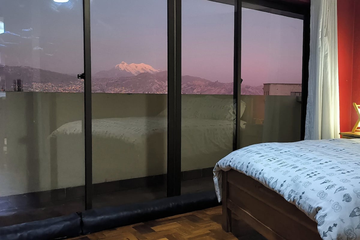 A bedroom with a bed, a nightstand, a lamp, and a view of a mountain through a sliding door.