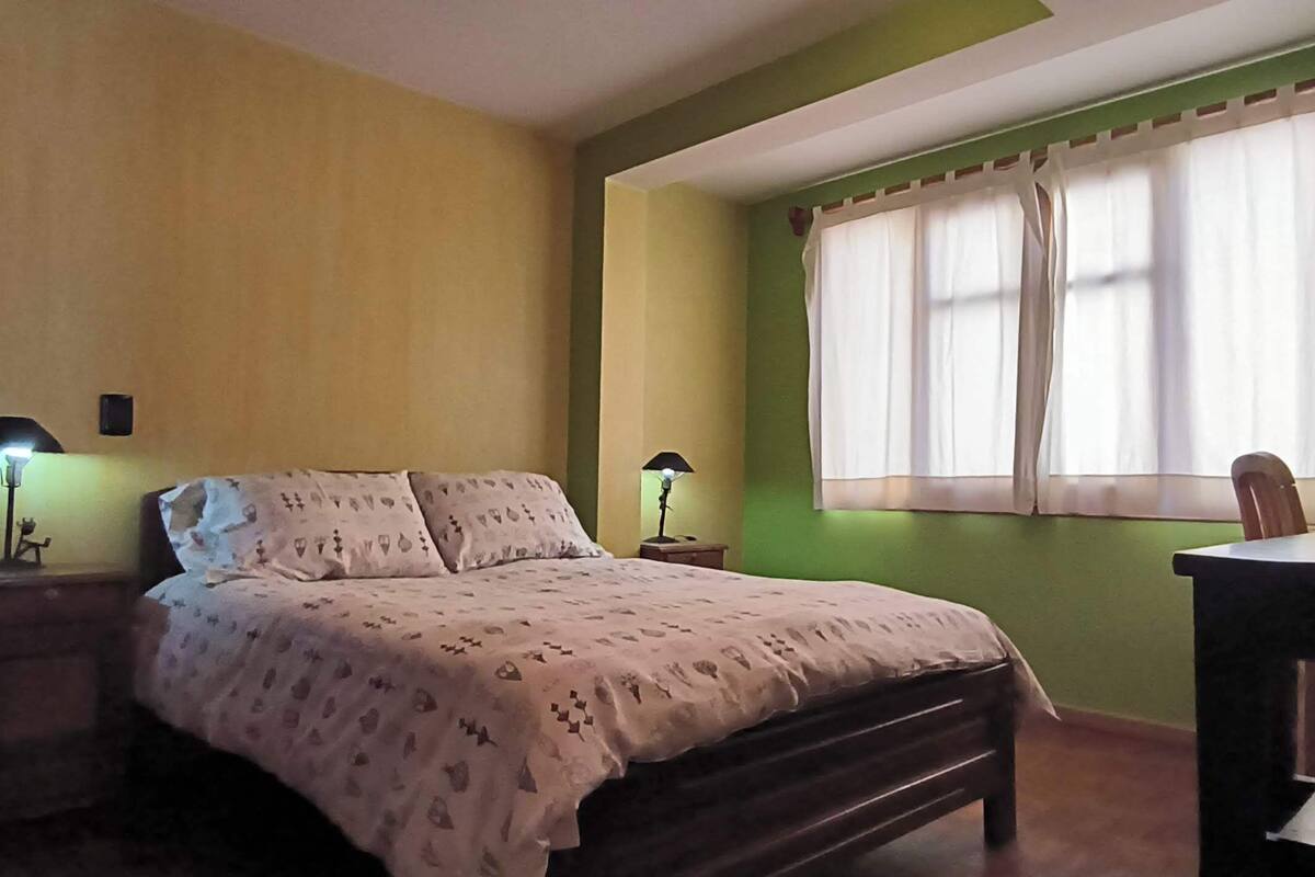 A bedroom with a bed, bedside lamps, a desk, and a window with curtains.