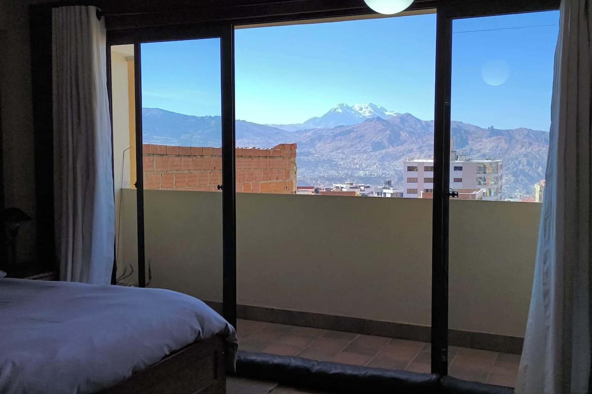 A bedroom with a bed, a view of mountains through a large window, and a balcony with a brick wall.