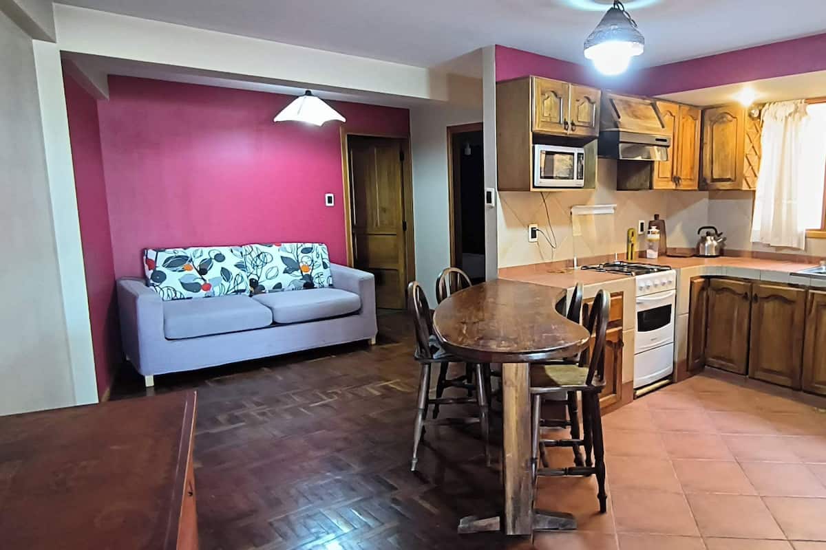 A compact kitchen with wooden cabinets, a dining table, and a sofa in a room with a tiled floor and a red accent wall.
