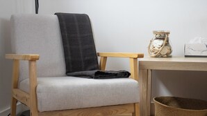 A wooden chair with a grey cushion and a dark grey blanket, a small woven basket, and a glass jar with a rope handle on a wooden table.
