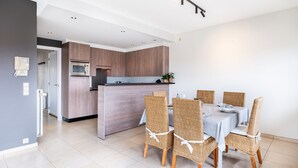 A modern kitchen with a central island, dining area with wicker chairs, and a visible door leading to another room. - Orangerie 202 Close to the Beach and Center (Koksijde)