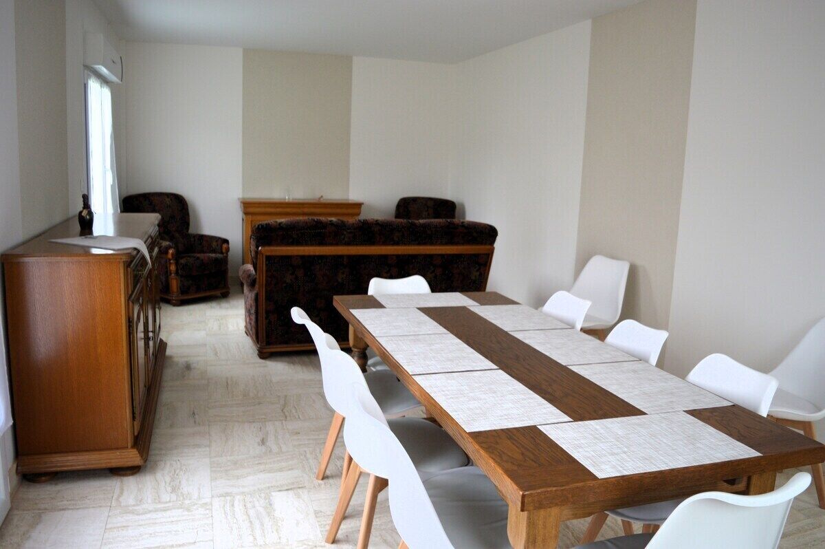 A dining area with a long wooden table, white chairs, and a dark patterned sofa.