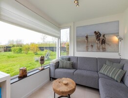 A modern living room with a grey sofa, a round wooden coffee table, and a large window offering a view of a garden and a painting of children by the sea.