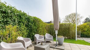 An outdoor patio with white plastic chairs, a woven table, and a grey umbrella. - Holiday Home in Nieuwvliet Near the Beach (Nieuwvliet)