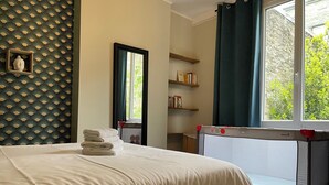 Un lit bien fait avec une serviette pliée, un berceau équipé de dispositifs de sécurité, une fenêtre avec des rideaux et une bibliothèque remplie de livres.
