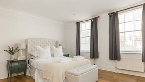 A bedroom with a large bed, a bedside table, a bench, and two windows with curtains. - Elegant 3BR Knightsbridge Residence near Harrods (London)