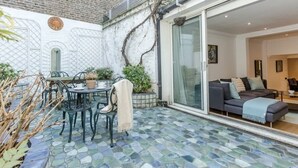 A patio with a mosaic floor, a dining table with chairs, a small potted plant, and a sliding glass door leading to an interior space with a sofa and a lamp. - Elegant 3BR Knightsbridge Residence near Harrods (London)