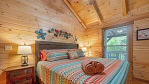 A cozy wooden cabin bedroom with a bed, two bedside lamps, a nightstand, and a window with blinds. - Creek Front Cabin in Pearisburg (Pearisburg)