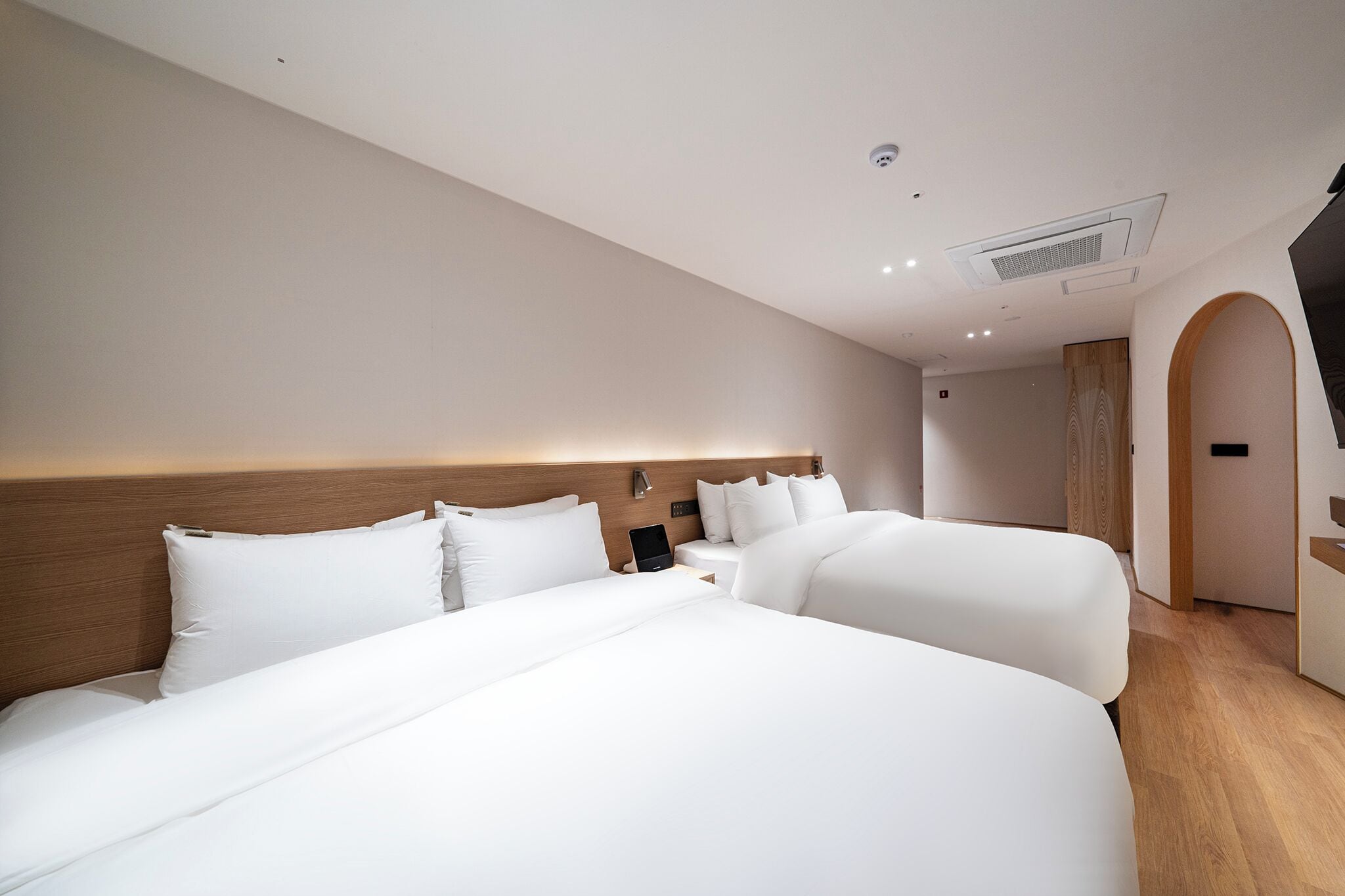 Una habitación de hotel moderna con dos camas grandes, un cabecero de madera, una mesita de noche con una lámpara y un equipo de aire acondicionado integrado.