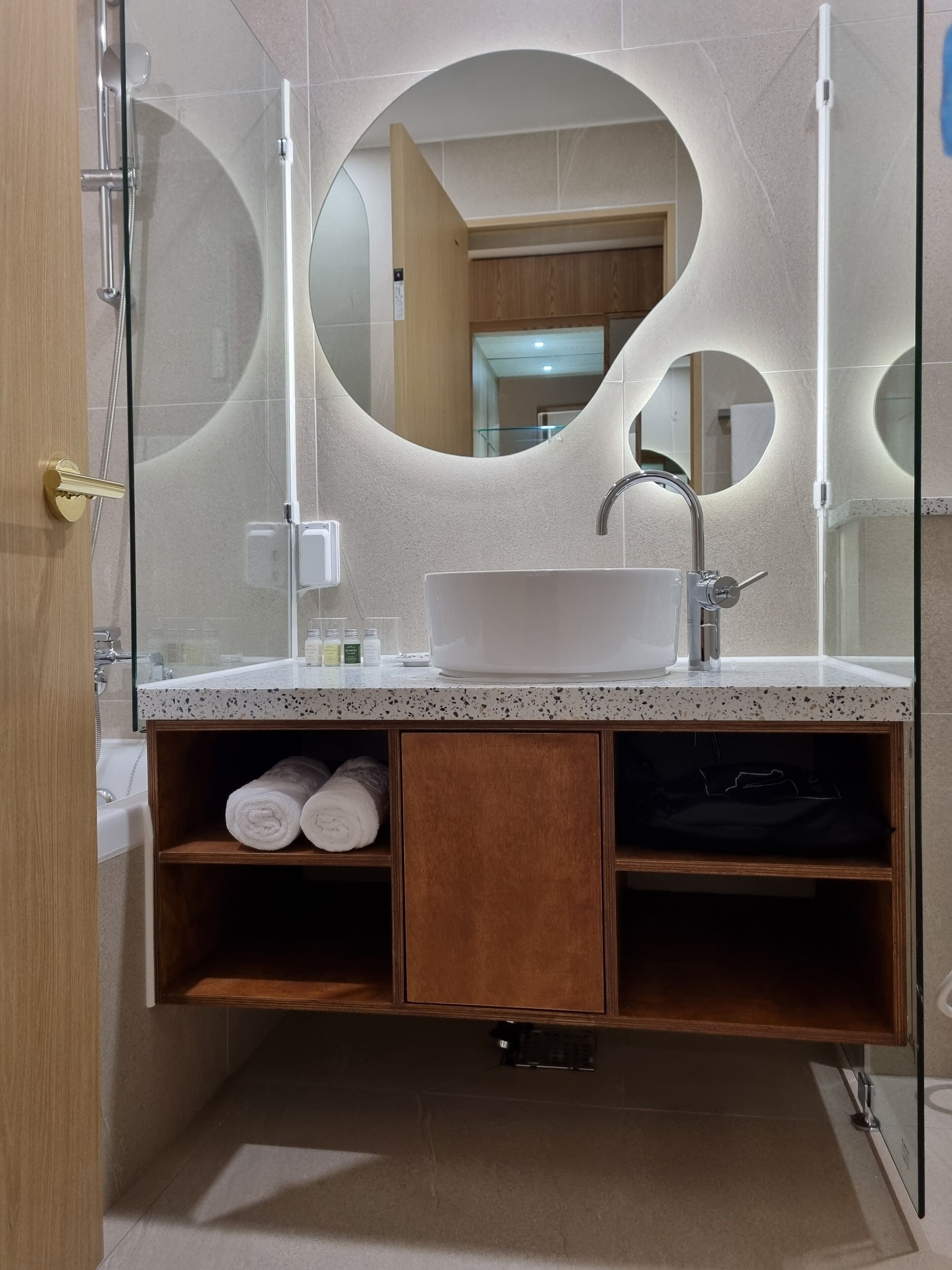 A modern bathroom with a freestanding bathtub, a large oval mirror, and a wooden vanity with shelves.