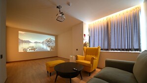 Una sala de estar moderna con una pantalla de proyector mostrando un paisaje pintoresco, un sillón amarillo, un sofá gris y una mesa de centro.