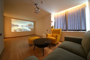 Una sala de estar moderna con una pantalla de proyector mostrando un paisaje pintoresco, un sillón amarillo, un sofá gris y una mesa de centro.