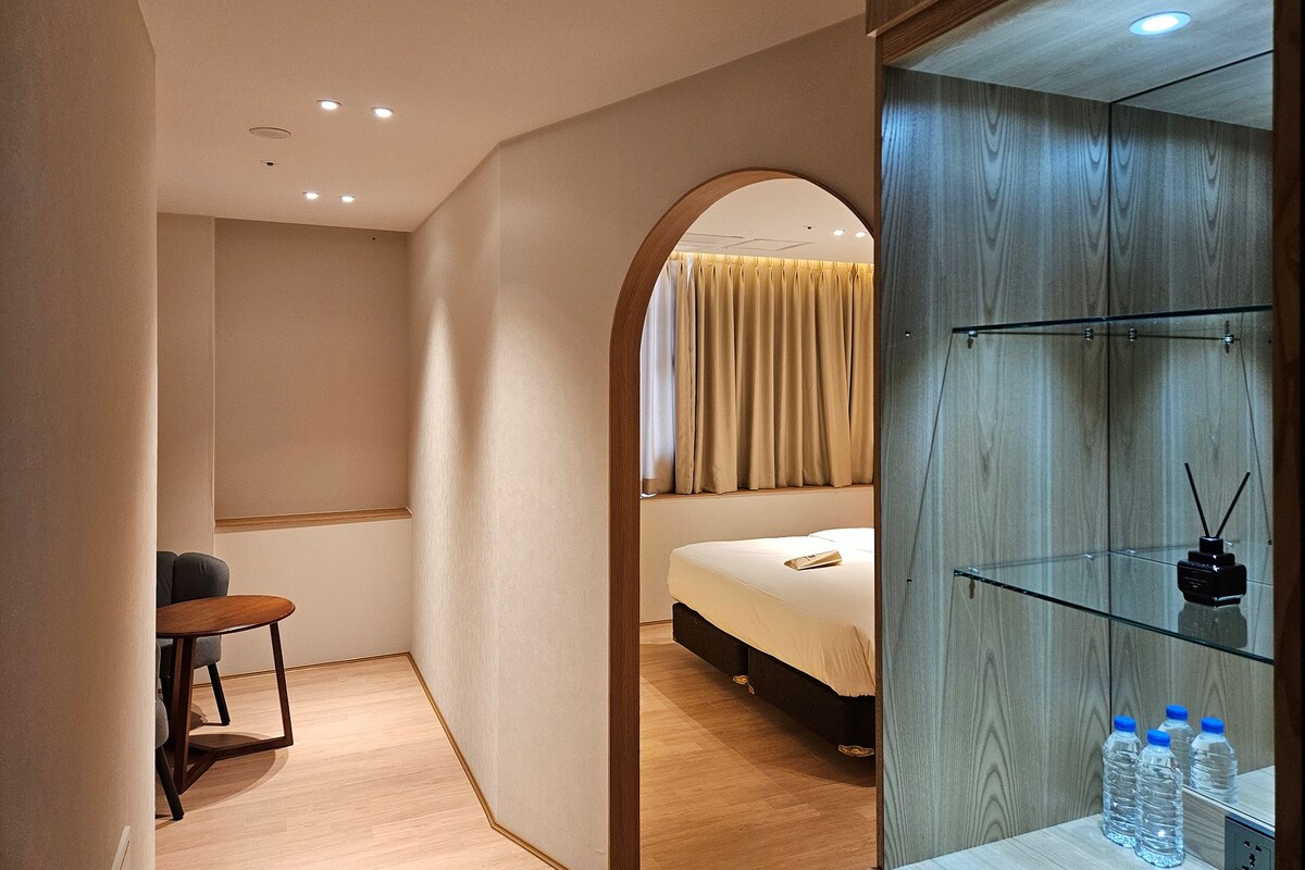 A modern hotel room with a bed, a small table, and a glass cabinet with water bottles.