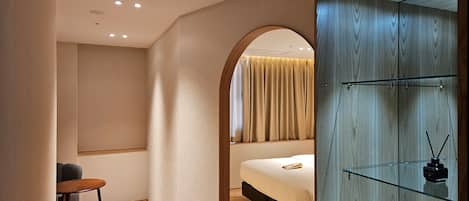 A modern hotel room with a bed, a small table, and a glass cabinet with water bottles.