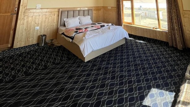 A bedroom with a wooden ceiling, a bed with a patterned bedspread, a window with a view, and a door.