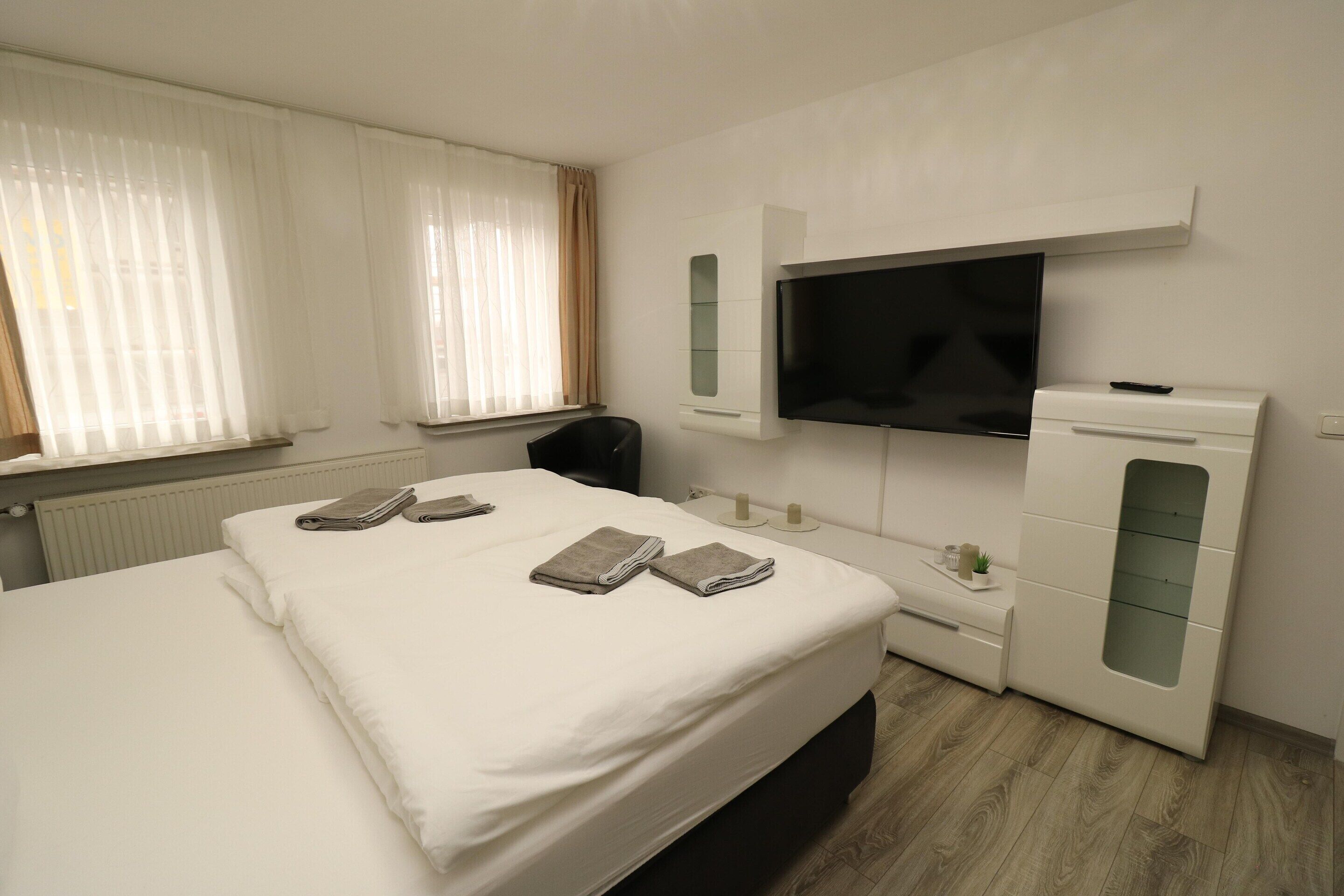 A hotel room with a large bed, a TV mounted on the wall, a desk with a chair, and a small table with a plant.