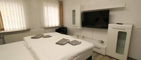 A hotel room with a large bed, a TV mounted on the wall, a desk with a chair, and a small table with a plant.