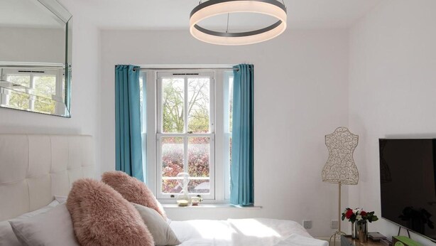 A modern bedroom with a bed, fluffy pillows, a large window with curtains, a pendant light, and a flat-screen TV.