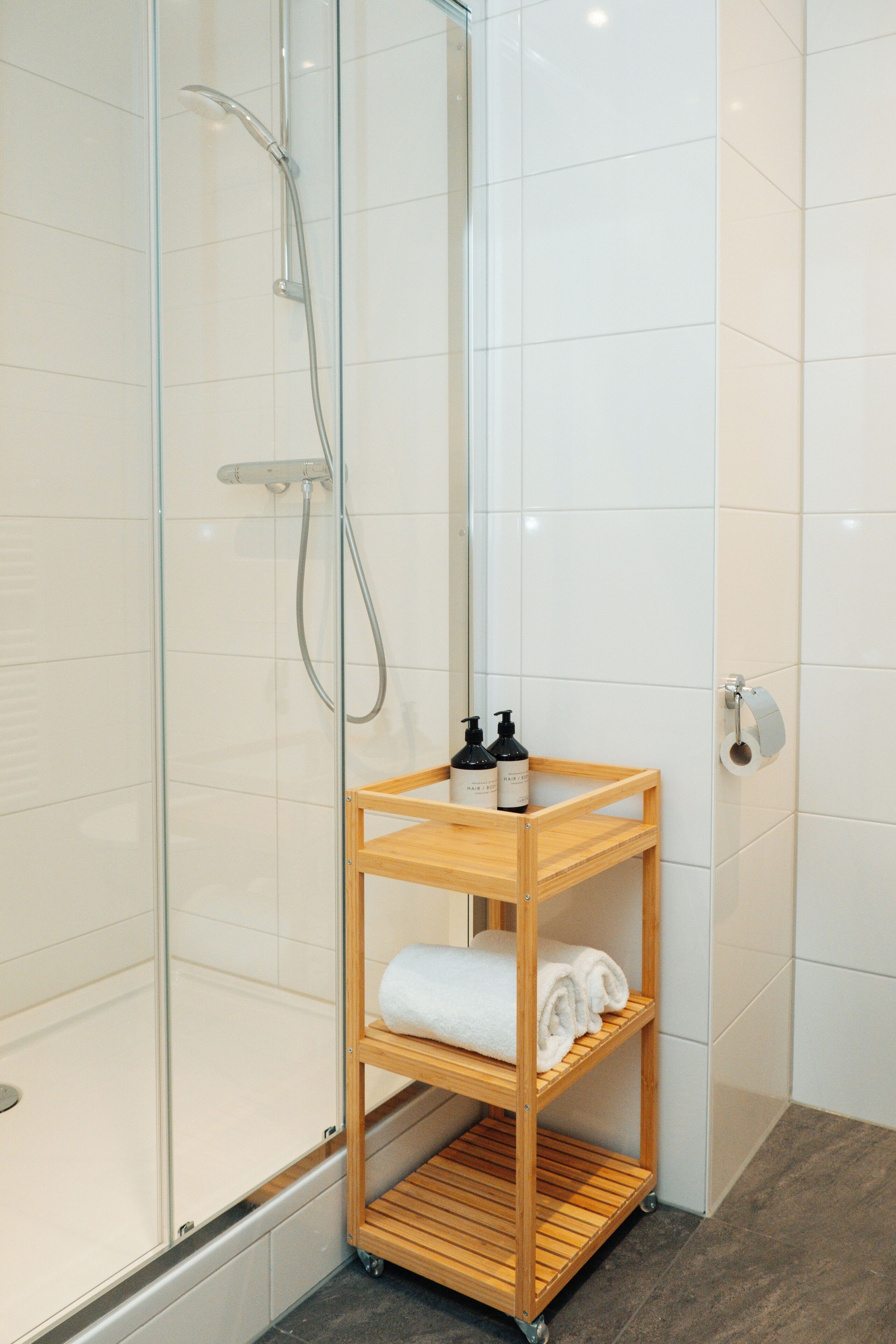 Une douche fermée par des parois en verre, dotée d’un pommeau de douche à main, d’une étagère en bois où sont posées des serviettes et des articles de toilette, et d’un porte-rouleau de papier toilette fixé au mur.