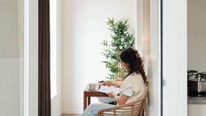 A woman sitting on a wicker chair in a narrow hallway with a plant, a small table, and a door leading to another room. - Urban Suites (Eindhoven)