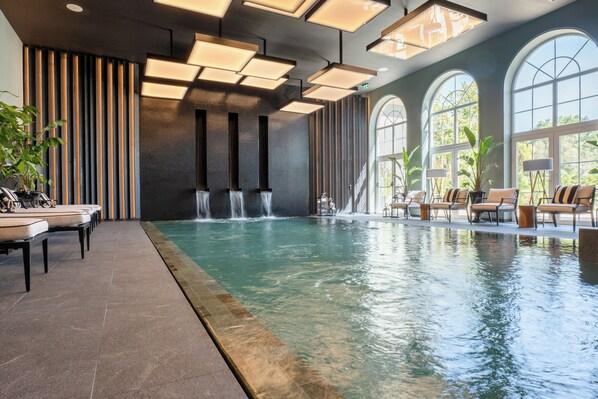 Una piscina interior con cascada, sillones de descanso y grandes ventanales en arco.