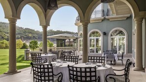An outdoor dining area with round tables and chairs, large windows, and a view of a green landscape. - Amoure Hotel (Vila Verde)