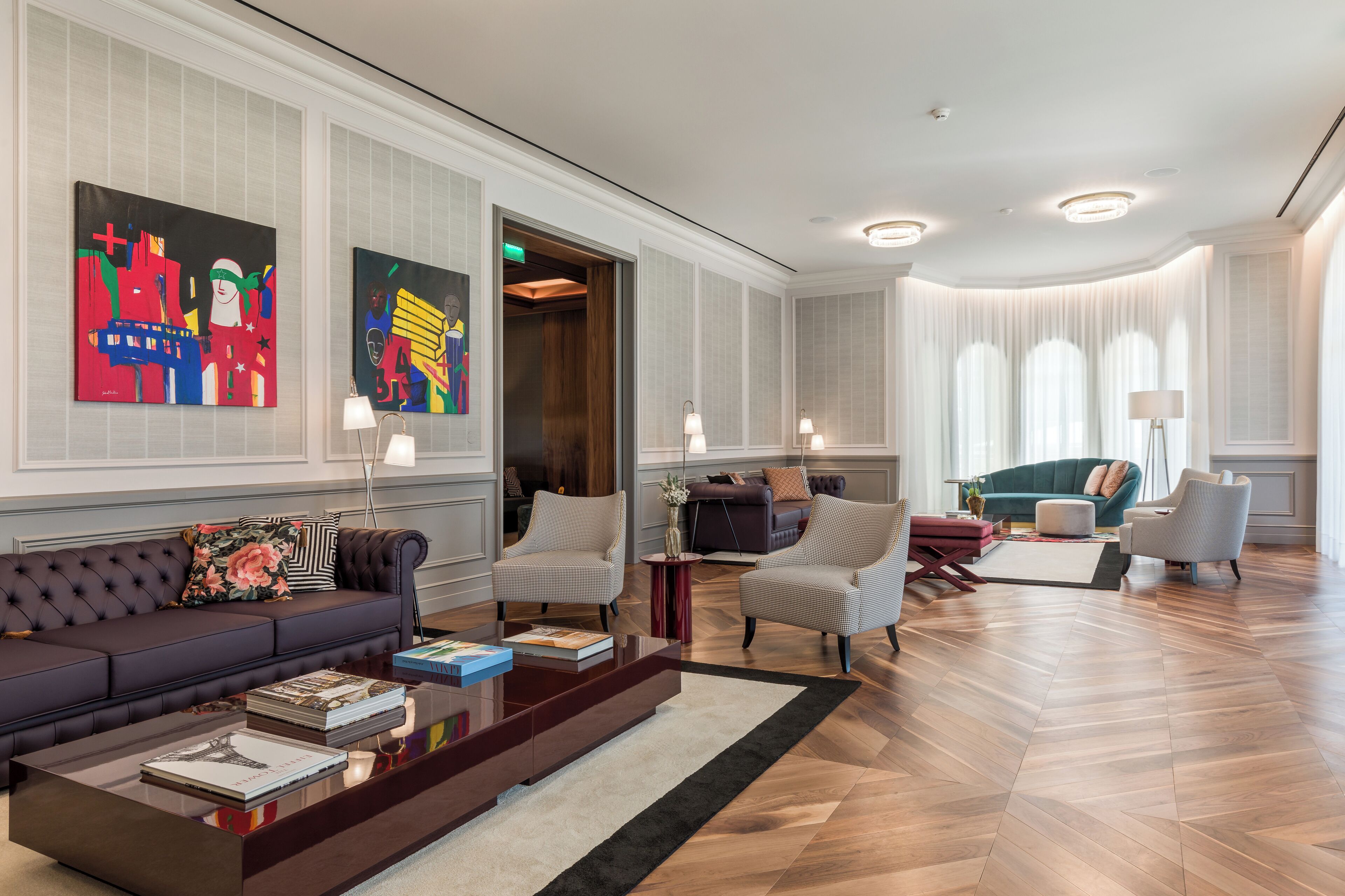 A spacious lobby with a dark brown sofa, two armchairs, a coffee table with books, and a large window with sheer curtains.