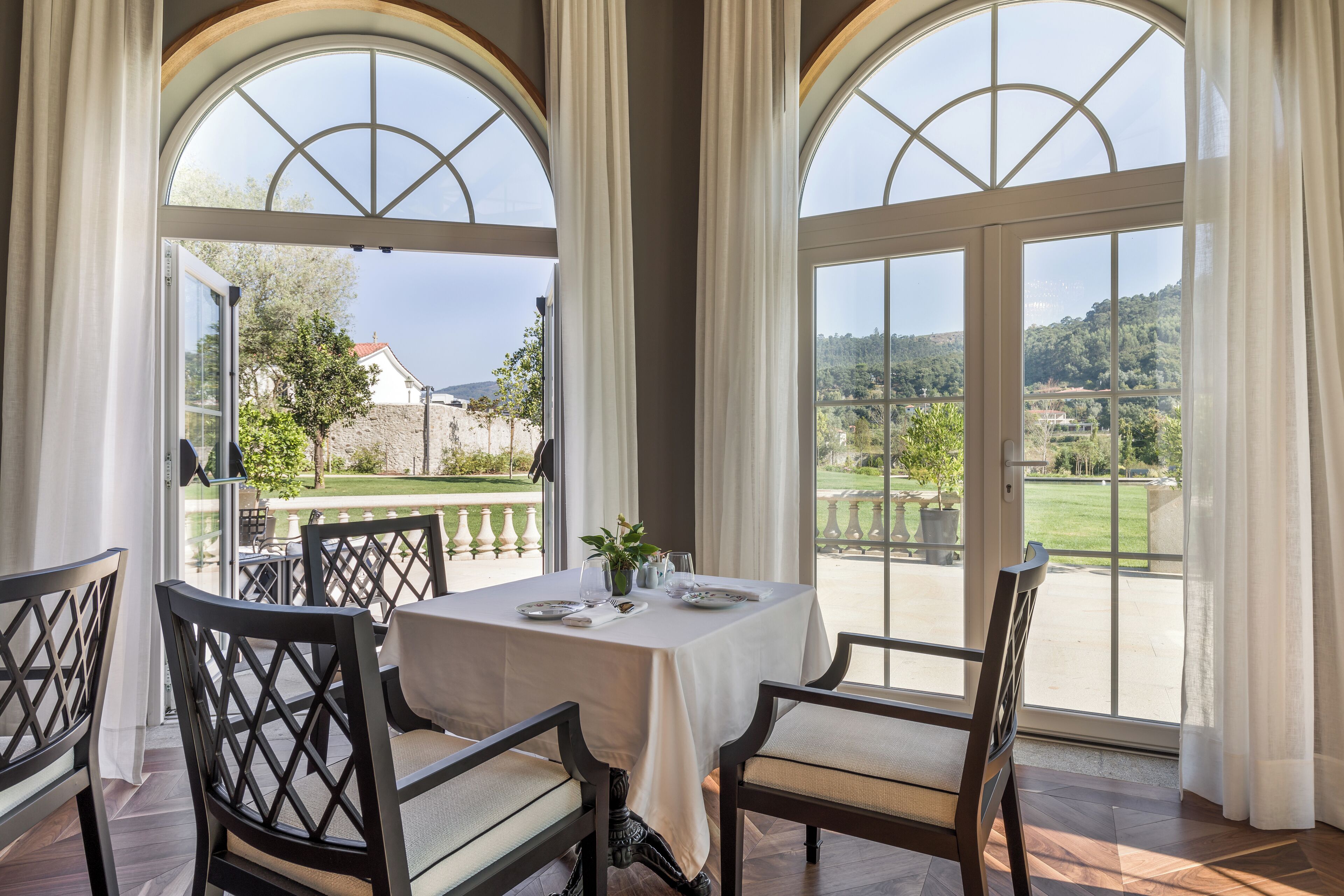 A dining area with a table set for four, large arched windows, and a view of a landscaped garden.